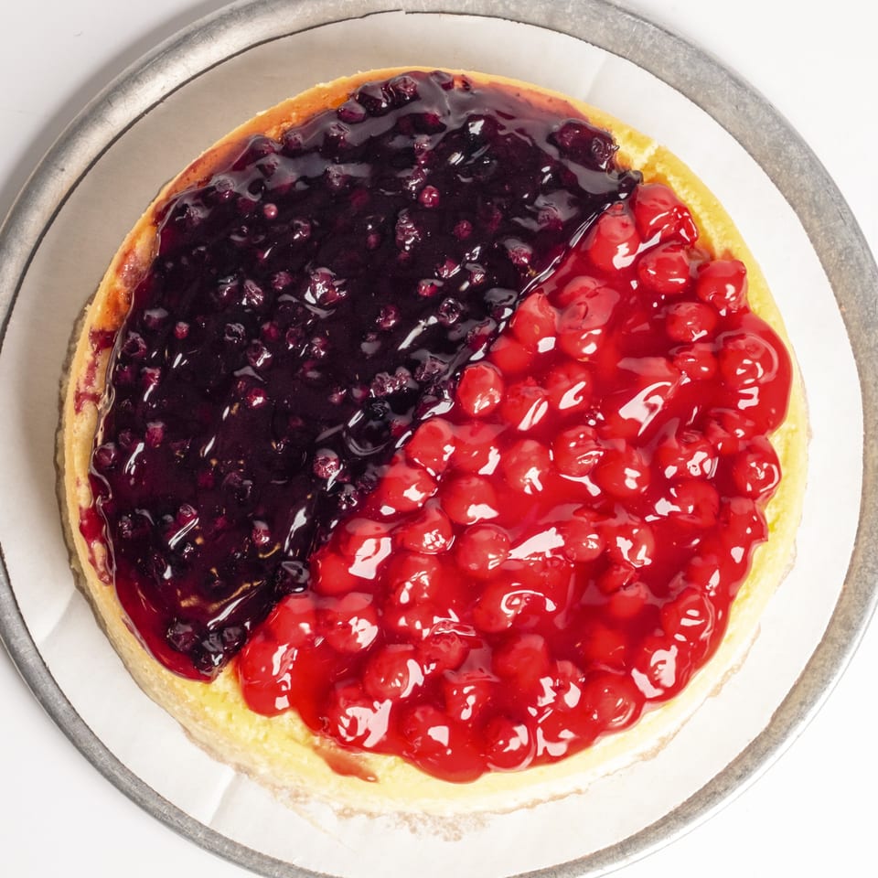 A whole cheesecake with half blueberry topping and half cherry topping on a metal tray.